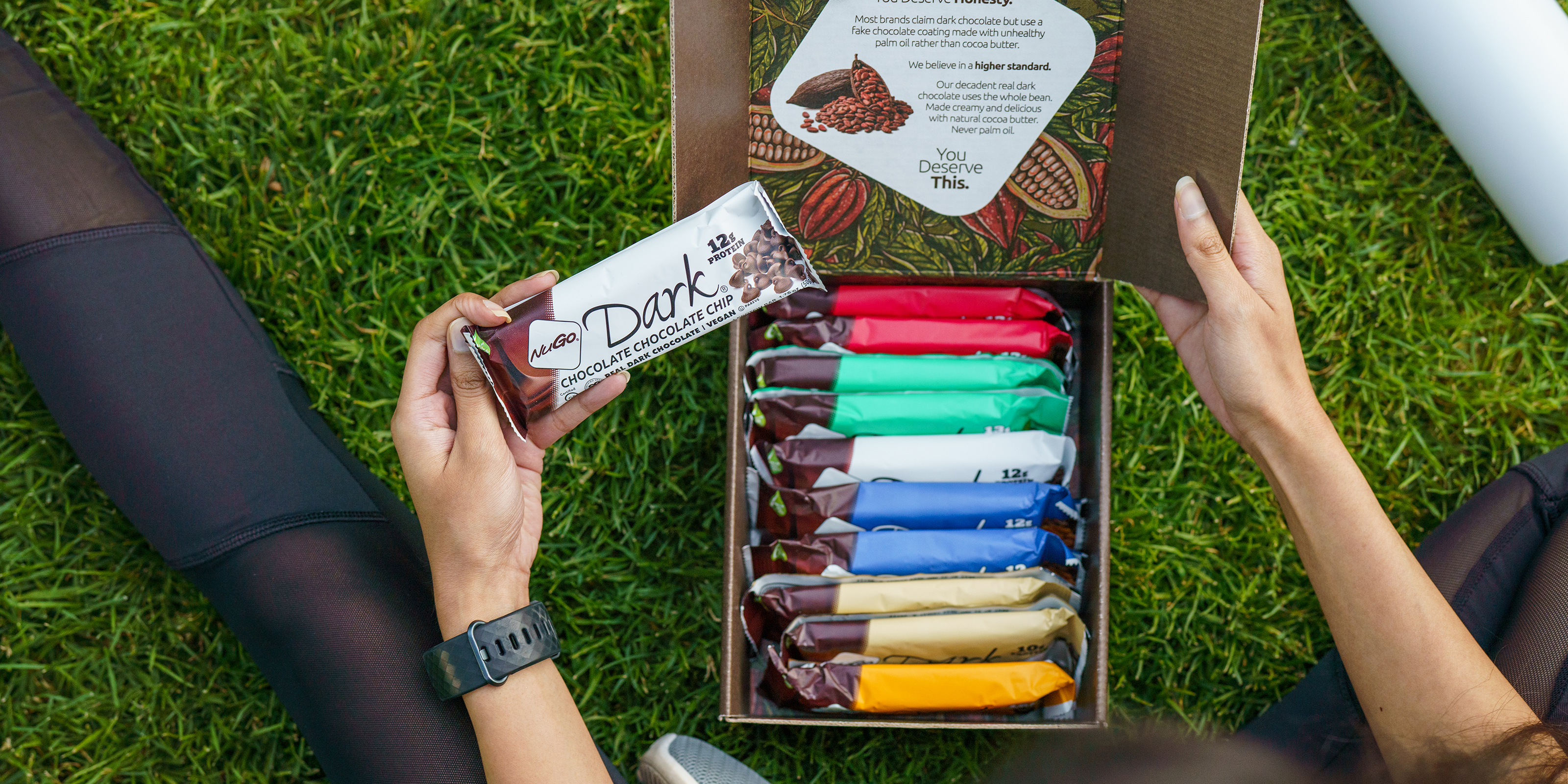 Woman sitting in grass with NuGo Dark Variety Pack box open and NuGo Dark Chocolate Chocollate Chip in hand