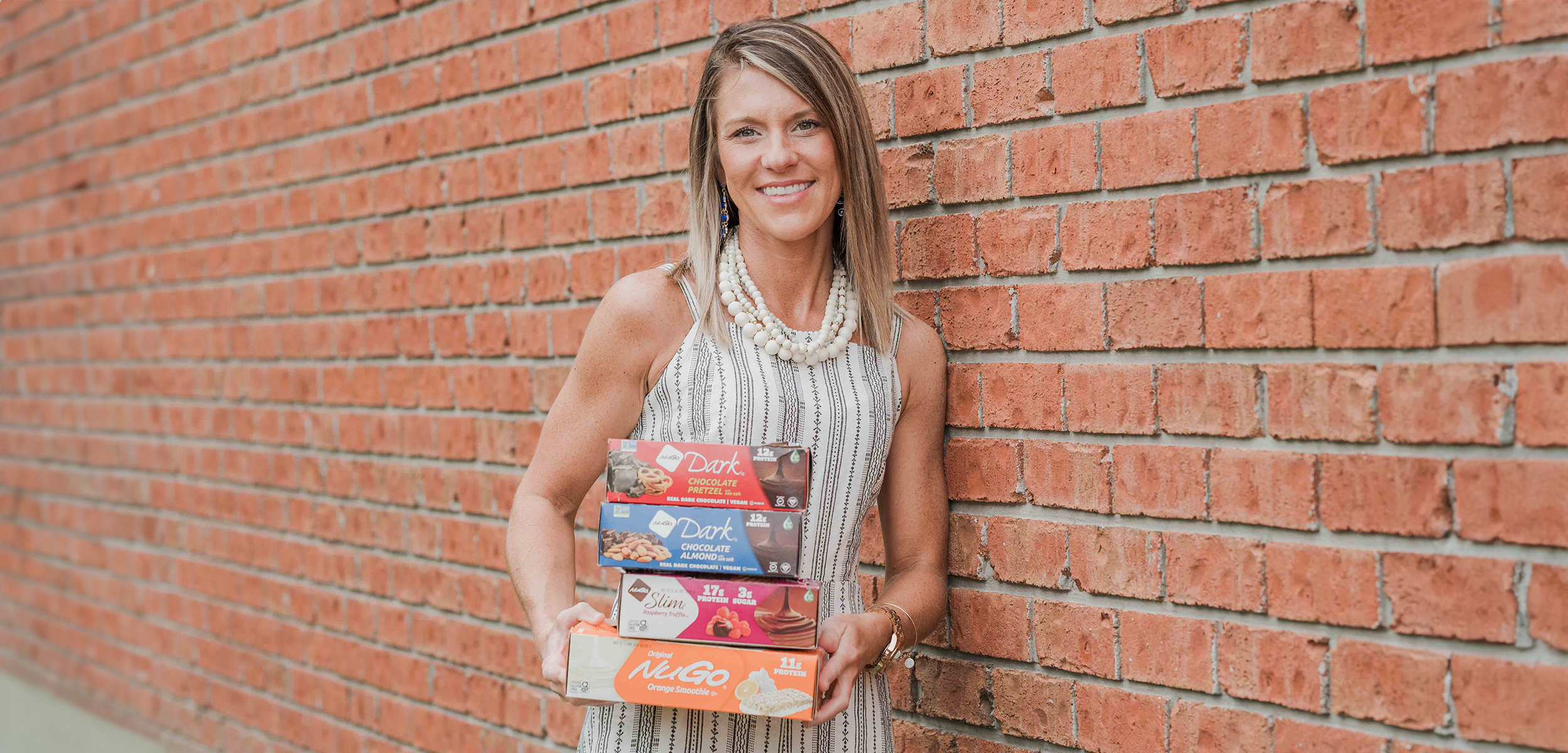 Woman hold 4 different NuGo boxes in front of Brick wall